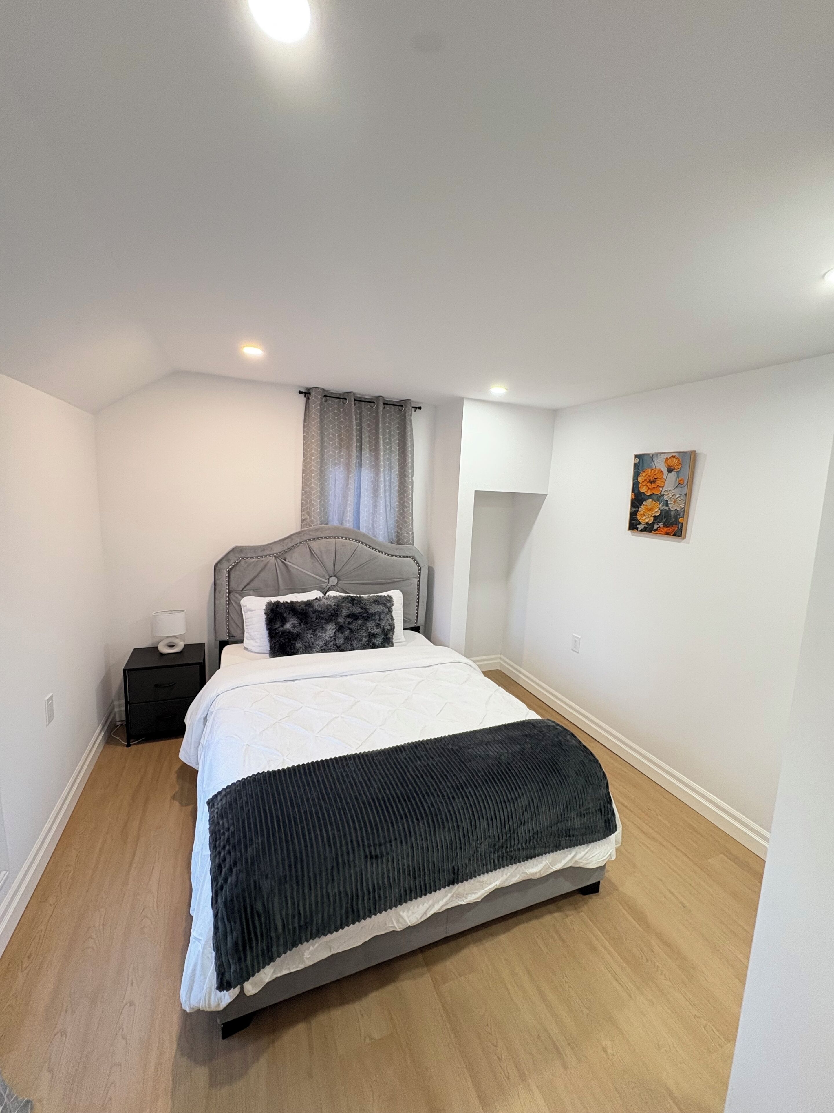 Quarto com cama, mesa de cabeceira, cabeceira, janela com cortinas e uma obra de arte na parede.