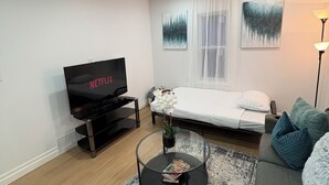 A modern living room with a flat-screen TV, a glass coffee table with books, a sofa, a bed, and two abstract paintings on the wall.