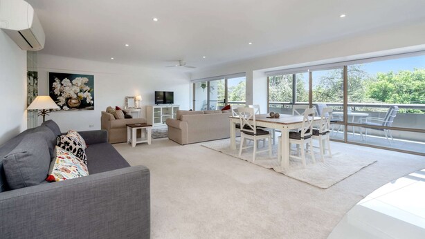 A spacious living area with a grey sofa, a beige sofa, a dining table with white chairs, and a large window offering a view of greenery. - Spacious 2-bed Apartment Near Dining & Attractions (Canberra)