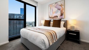 A modern bedroom with a large bed, a city view, a painting on the wall, and a nightstand with a lamp. - Southbank Stay in Prima Tower | Premium Facilities (Southbank)