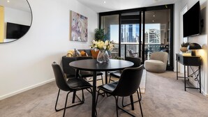A modern living room with a round table, black chairs, a sofa, a TV, and a large window with a city view. - Southbank Stay in Prima Tower | Premium Facilities (Southbank)