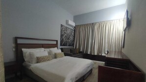 Habitación de hotel con una cama grande, una mesita de noche, un televisor montado en la pared y un ventanal con cortinas.