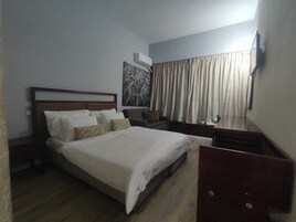 Habitación de hotel con una cama grande, una mesita de noche, un televisor montado en la pared y un ventanal con cortinas.