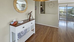 A modern living room with a wooden floor, a white console table with a telescope, a round mirror, a hanging light fixture, and a view of the outdoors through a glass door. - Coastal Upper-level Home With Pool Access (Batehaven)