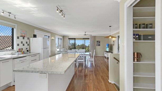 A modern kitchen with a marble island, wooden flooring, and a dining area with white furniture. - Coastal Upper-level Home With Pool Access (Batehaven)