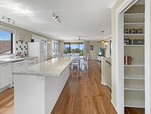 A modern kitchen with a marble island, wooden flooring, and a dining area with white furniture. - Coastal Upper-level Home With Pool Access (Batehaven)