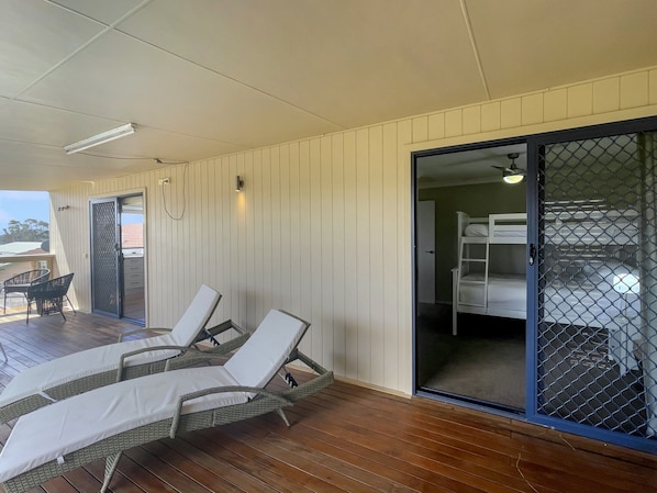 Une pièce avec une terrasse en bois, deux fauteuils de relaxation, une porte-fenêtre coulissante donnant accès à une chambre avec des lits superposés, et une petite table avec des chaises.