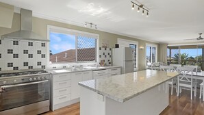 A modern kitchen with a central island, stainless steel appliances, and large windows offering a view of the outdoors. - Coastal Upper-level Home With Pool Access (Batehaven)