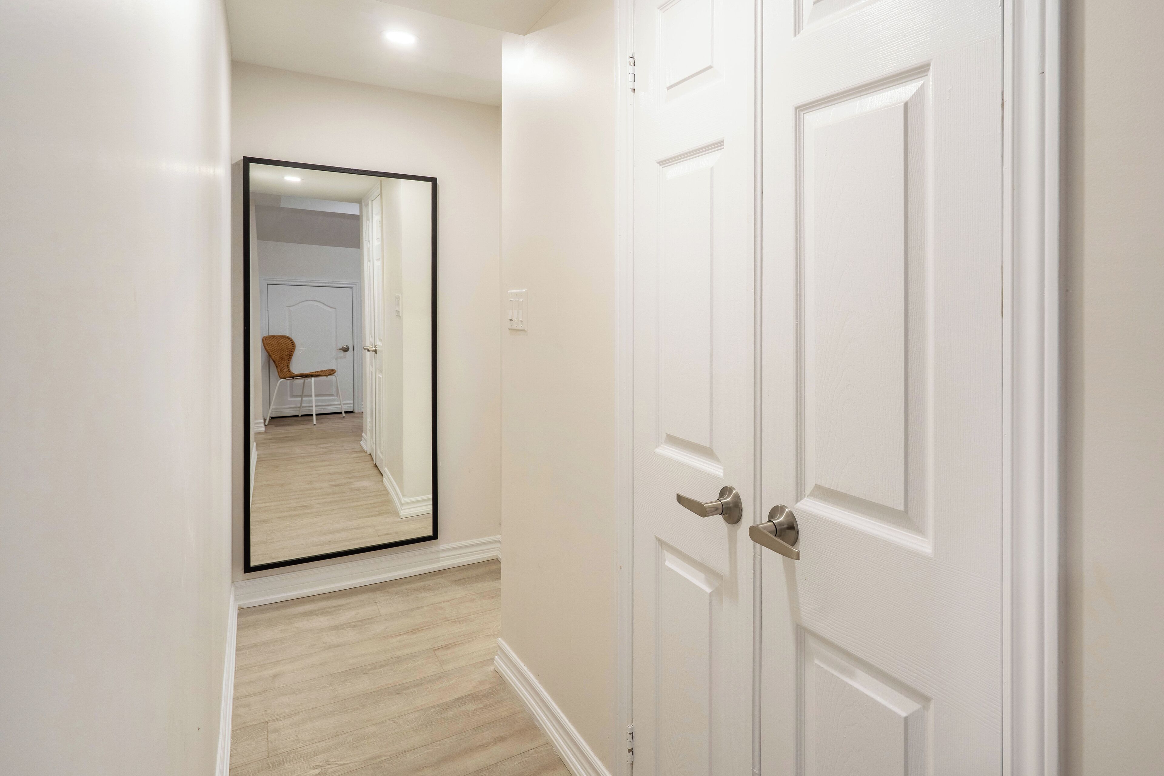 Un pasillo con una puerta blanca, suelo de madera y un espejo que refleja una habitación con una silla y una puerta.