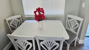 Un coin repas avec une table et des chaises blanches, un vase rempli de fleurs rouges et une fenêtre avec des stores.