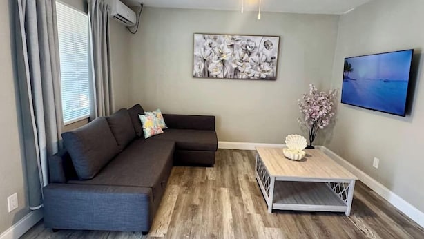 Un salon avec un canapé d’angle gris foncé, un ventilateur de plafond, un téléviseur fixé au mur et une table basse en bois.