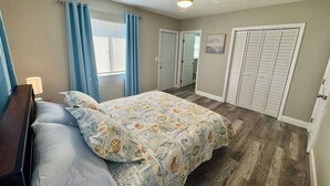Une chambre à coucher comprenant un lit, un ventilateur de plafond, une fenêtre avec des rideaux et une porte donnant sur une autre pièce.