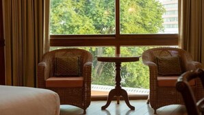 A room with two wicker chairs, a small round table, and a large window with a view of trees and buildings. - Sailom Hotel Hua Hin (Hua Hin)