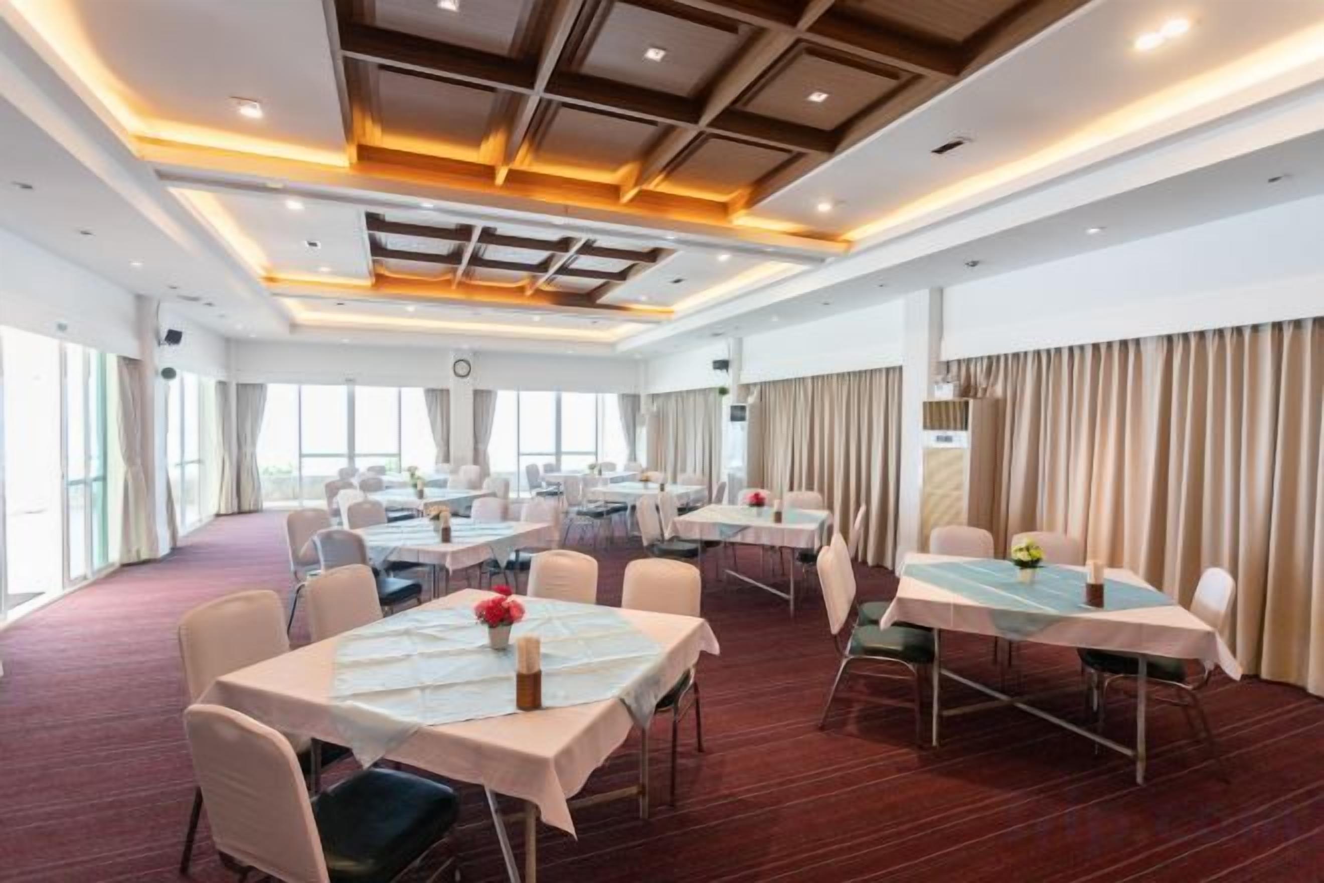 A hotel conference room with multiple tables, chairs, and a large window providing natural light.