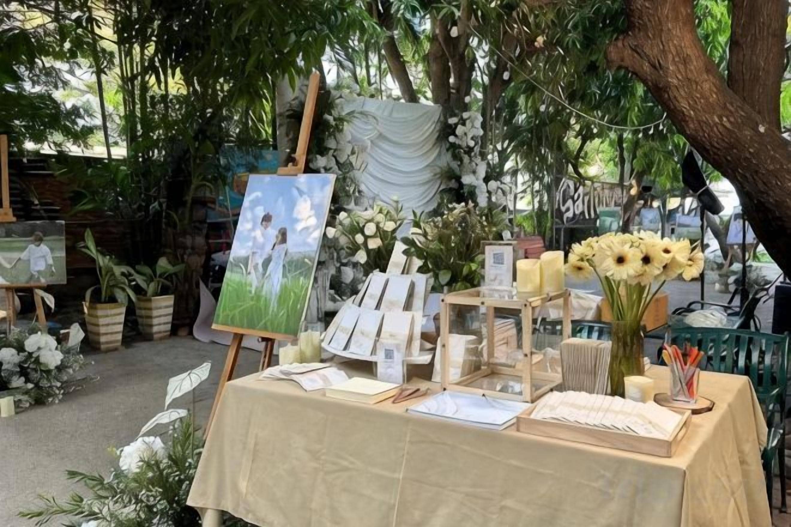 An outdoor art display with paintings, a table with books and candles, and floral arrangements.