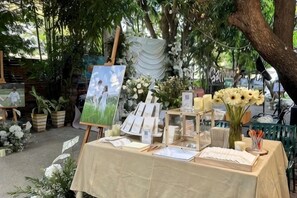 Une exposition d’art en plein air avec des tableaux, une table ornée de livres et de bougies, et des compositions florales.