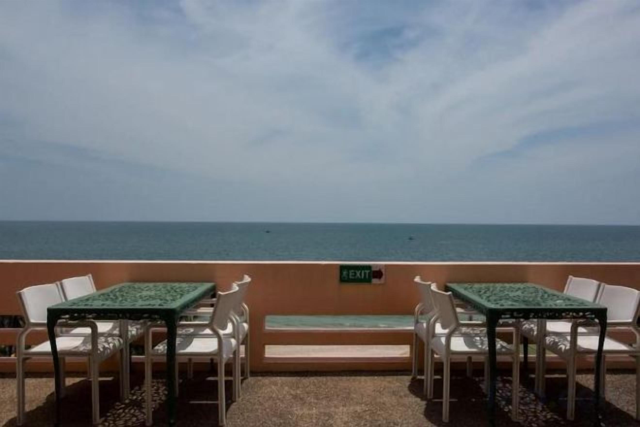 An outdoor seating area with tables and chairs facing the sea, a sign indicating an exit, and a clear sky.