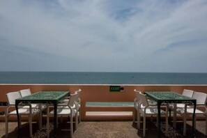 Een terras met tafels en stoelen gericht op de zee, een bord met een aanduiding voor een uitgang, en een heldere lucht.