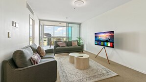 Ein modernes Wohnzimmer mit grauem Sofa, einem Couchtisch, einem Flachbildfernseher auf einem Standfuß und einem großen Fenster mit Blick auf die Stadt.