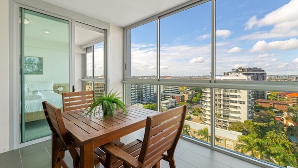 Ein Balkon mit Holztisch und -stühlen, einem Blumentopf und Blick auf die Stadt durch große Fenster.
