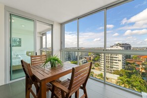 Varanda com mesa e cadeiras de madeira, planta em vaso e vista da cidade por grandes janelas.