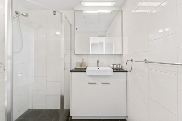 A modern bathroom with a glass shower enclosure, a white sink with a mirror above it, and a towel rack on the wall. - Kangaroo Point 2-bed Flat With Gym & Pool (Brisbane)