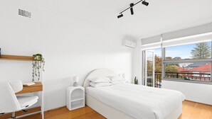 A modern bedroom with a wooden floor, a white bed, a desk with a chair, and a large window with a view of buildings and trees. - Bright 2-bed Flat With Balcony in Vaucluse (Sydney)