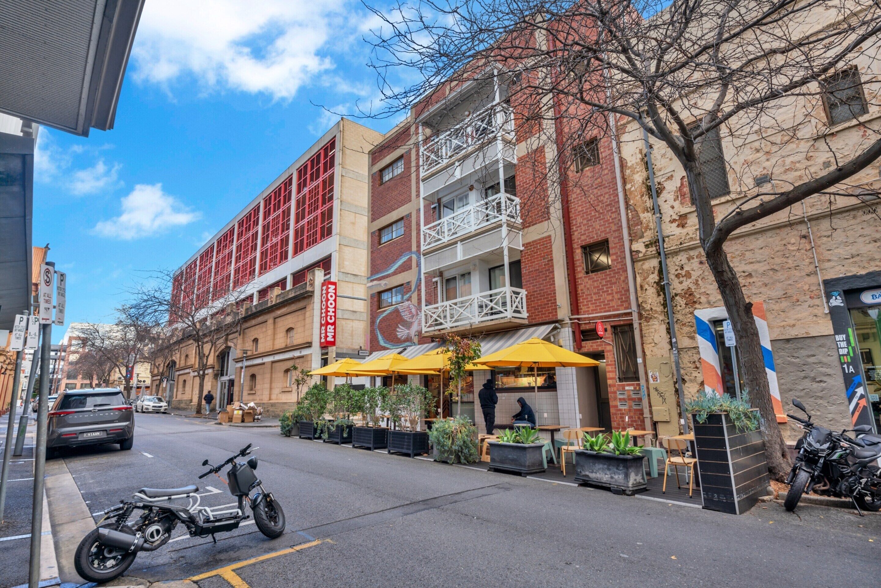A street view with a building featuring outdoor seating, a motorcycle parked on the sidewalk, and a tree with bare branches.