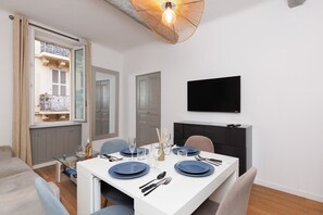 A dining area with a white table set for four, a television mounted on the wall, and a view of a balcony with a wrought iron railing. - Upstays Banane Palais 27m² Apartment (Cannes)