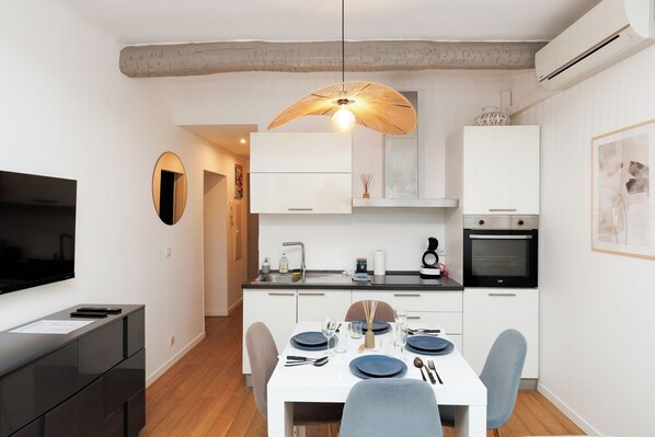 A modern kitchen with white cabinets, a dining table set for four, and a TV on the left. - Upstays Banane Palais 27m² Apartment (Cannes)
