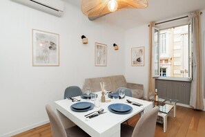 A dining area with a white table set for four, a sofa, and a window with curtains. - Upstays Banane Palais 27m² Apartment (Cannes)