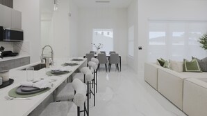 A modern kitchen with a long countertop, a dining area with a white table and chairs, and a sofa with cushions.
