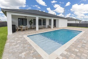 A rectangular swimming pool with a tiled deck, a covered patio area with seating, and a white building with a dark roof. - Is This Cape`s Best Secret? | 4BR Midnight Pool (Cape Coral)