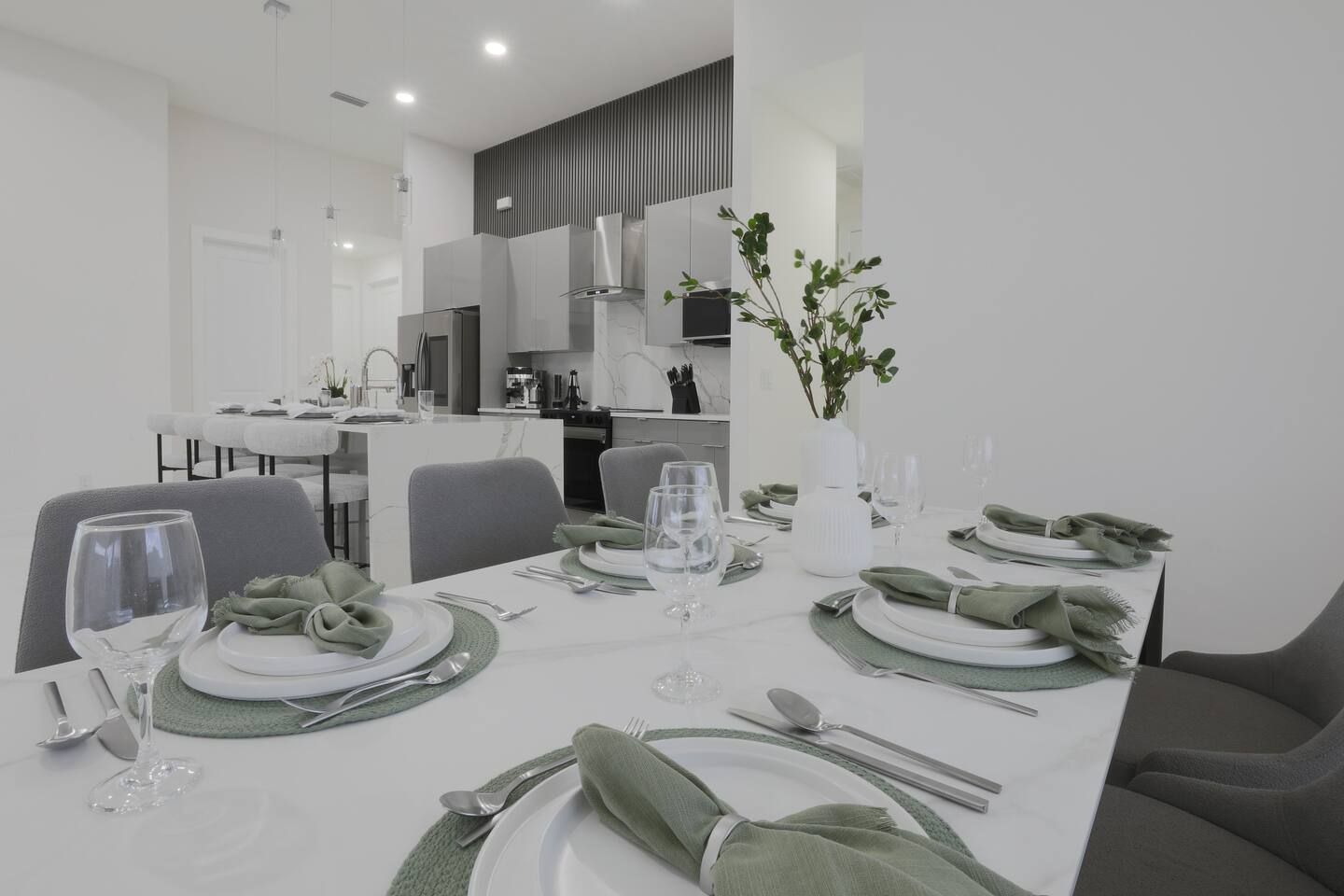 Un'area pranzo moderna con un tavolo bianco apparecchiato per un pasto, sedie grigie e una cucina sullo sfondo.