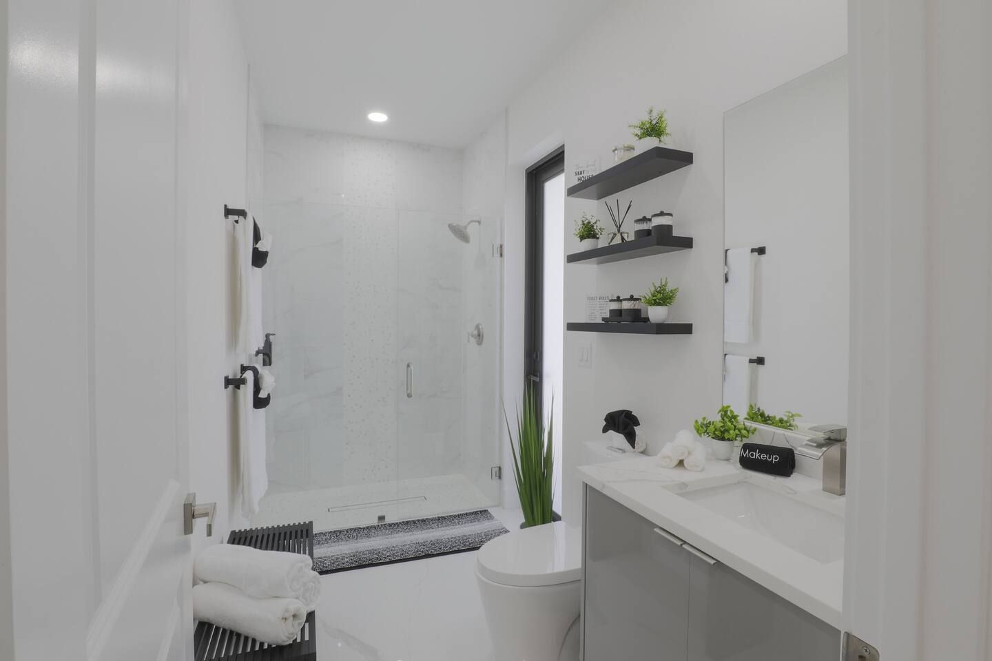A modern bathroom with a shower, toilet, and sink. There are shelves with plants and toiletries.