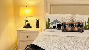 Un lit bien fait, avec une literie à motifs floraux, une lampe de chevet et des coussins décoratifs, dont un avec un motif représentant un ours et une montagne.