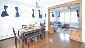 Un salon spacieux avec une table à manger en bois, des chaises et un canapé. On y trouve un ventilateur de plafond, un lustre et une fenêtre avec des rideaux.