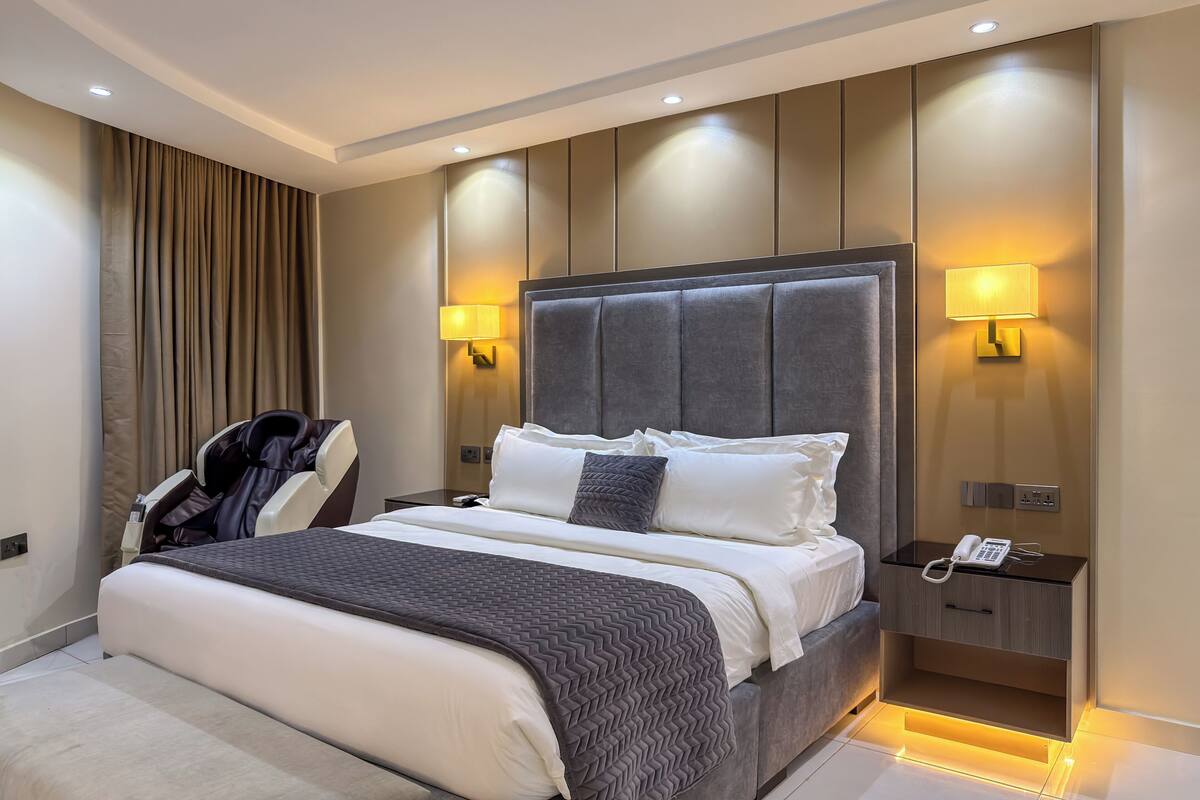 A modern hotel room with a large bed, two bedside lamps, a phone on the nightstand, and a massage chair.