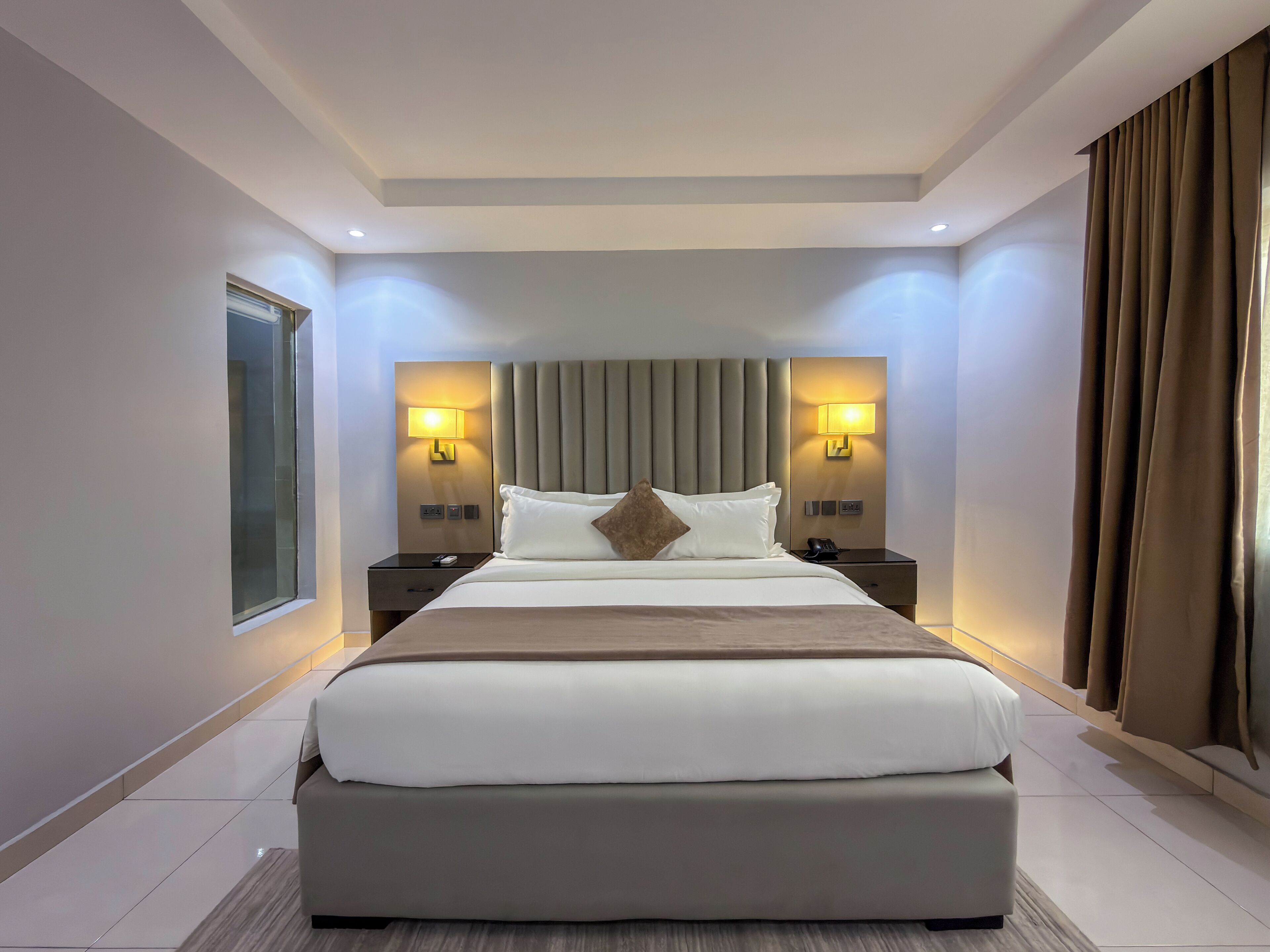 A modern hotel room with a large bed, two bedside tables with lamps, a window, and a curtain.