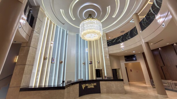 A hotel lobby with a modern design, featuring a central chandelier, a reception desk, and a high ceiling with circular lighting.
