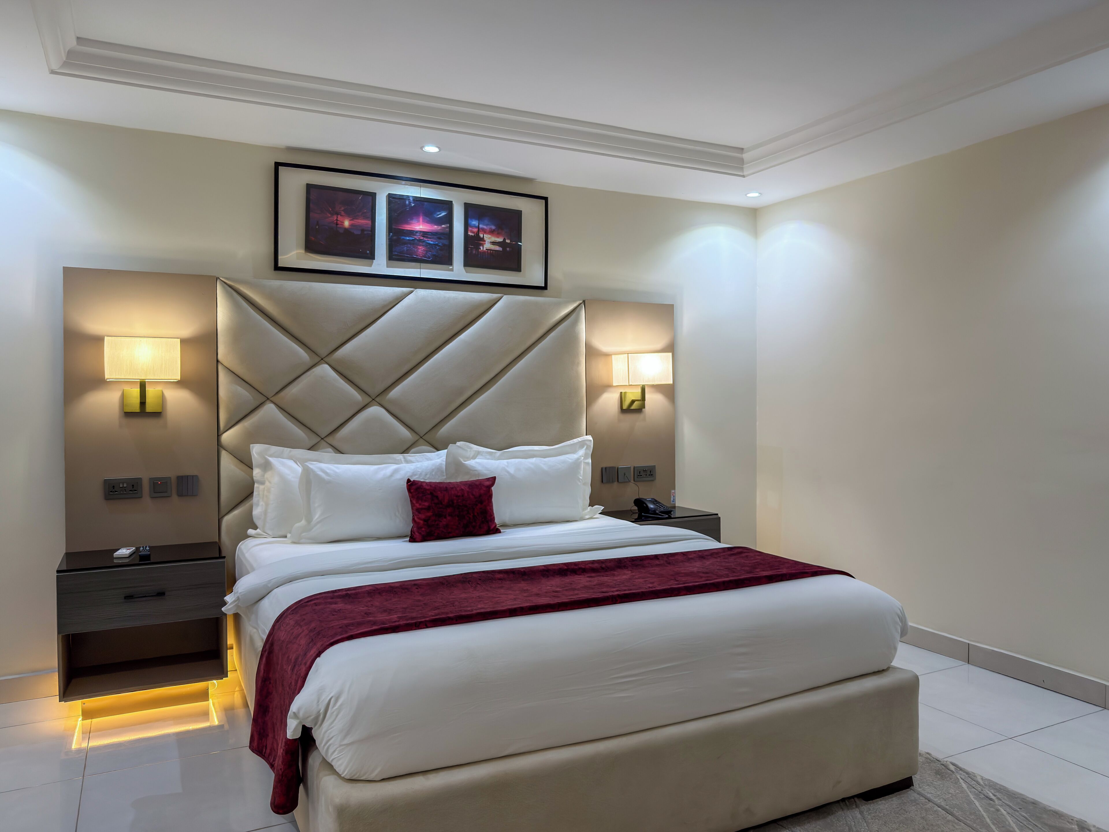 A modern hotel room with a large bed, a headboard with a diamond pattern, bedside lamps, a nightstand, and a wall-mounted phone.