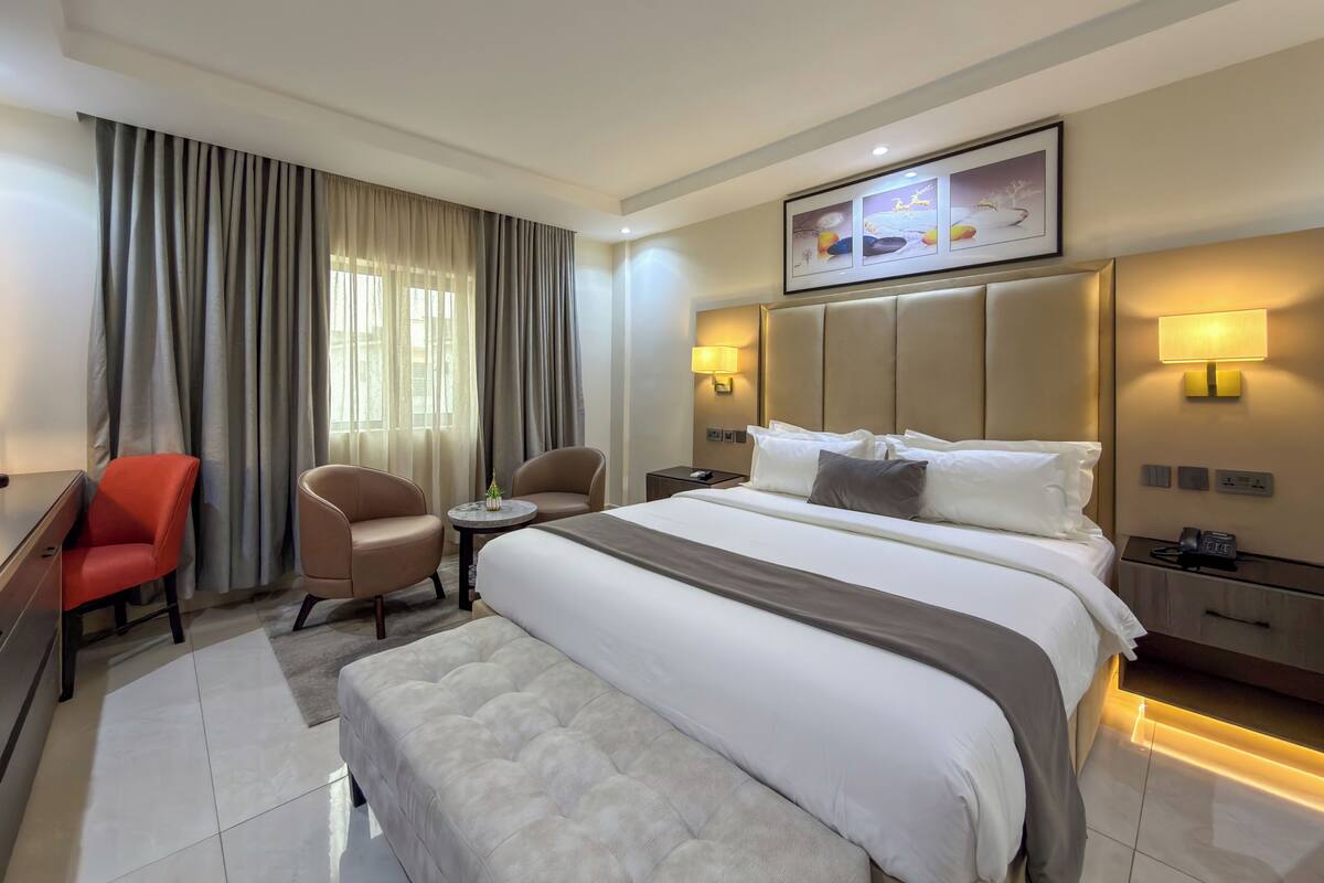 A hotel room with a bed, two chairs, a desk, a lamp, and a painting on the wall.
