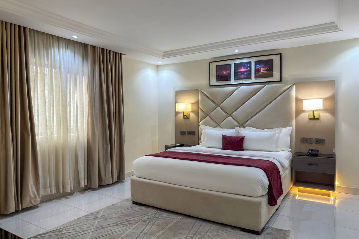 A hotel room with a large bed, two bedside tables, a headboard with a diamond pattern, and a window with curtains.