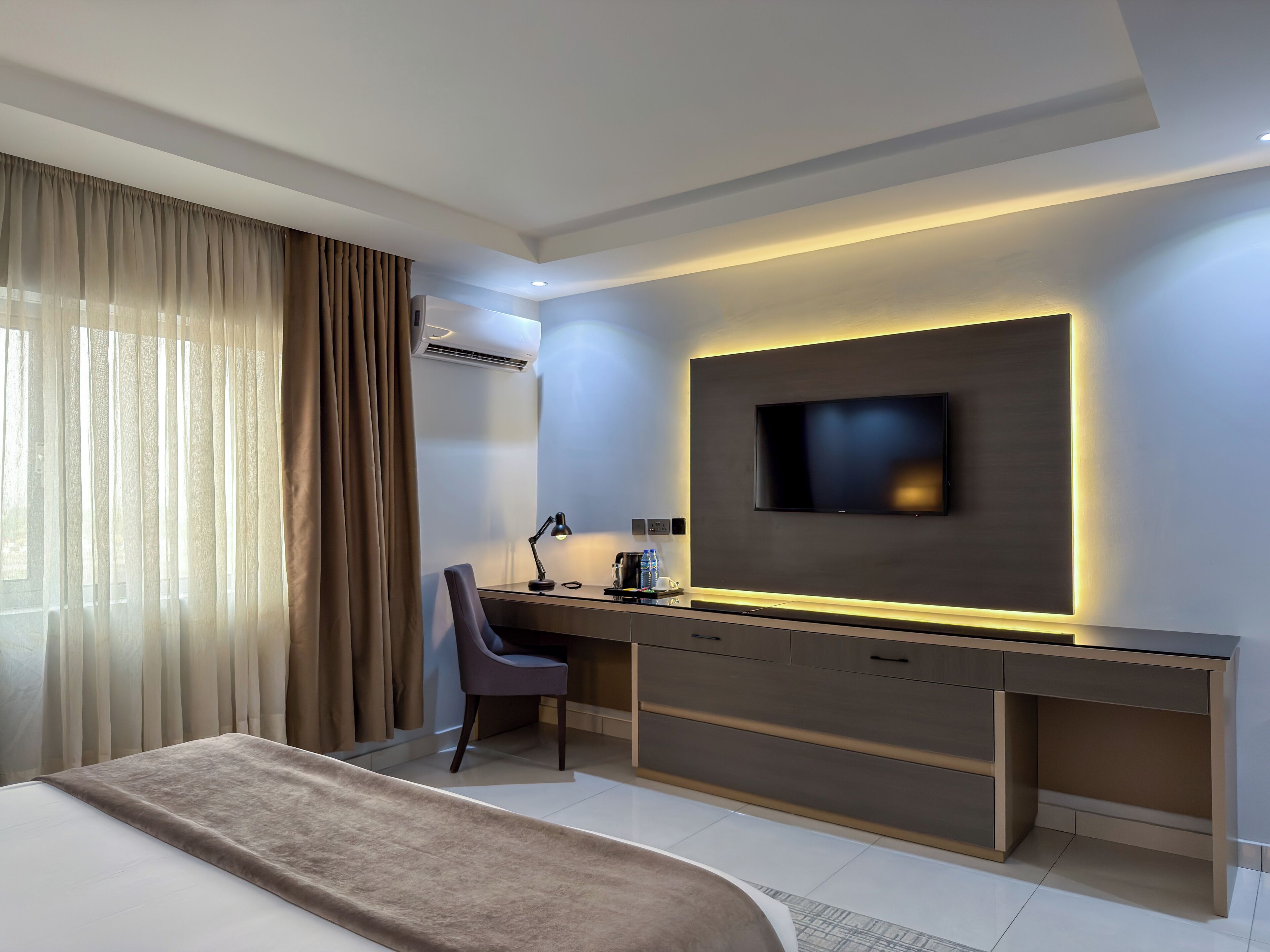 A modern hotel room with a bed, a desk with a chair, a flat-screen TV, and an air conditioning unit.