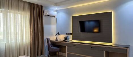 A modern hotel room with a bed, a desk with a chair, a flat-screen TV, and an air conditioning unit.