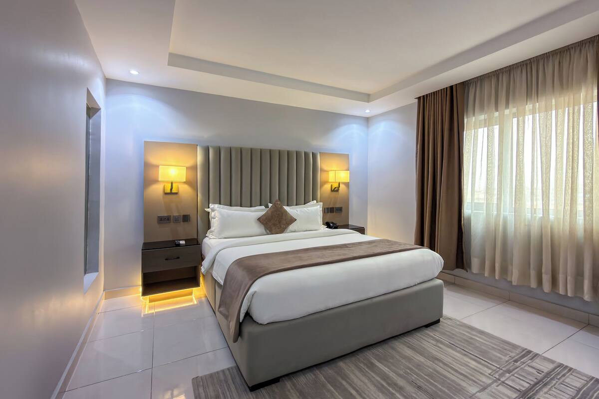 A modern hotel room with a large bed, bedside lamps, a nightstand, and a view of the outdoors through sheer curtains.