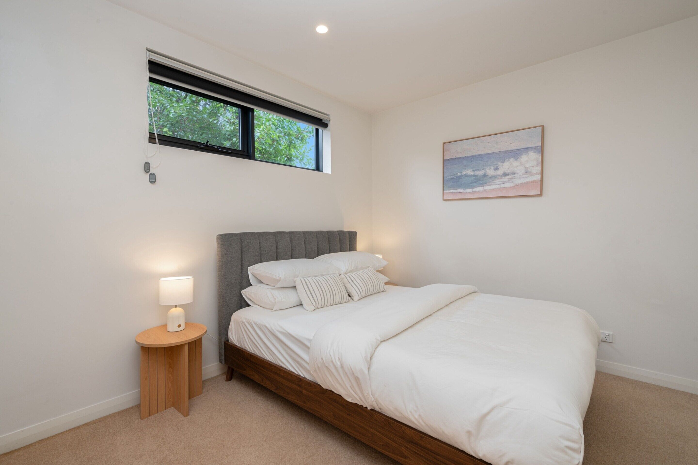 A bedroom with a bed, a bedside table with a lamp, a window with blinds, and a wall-mounted picture.
