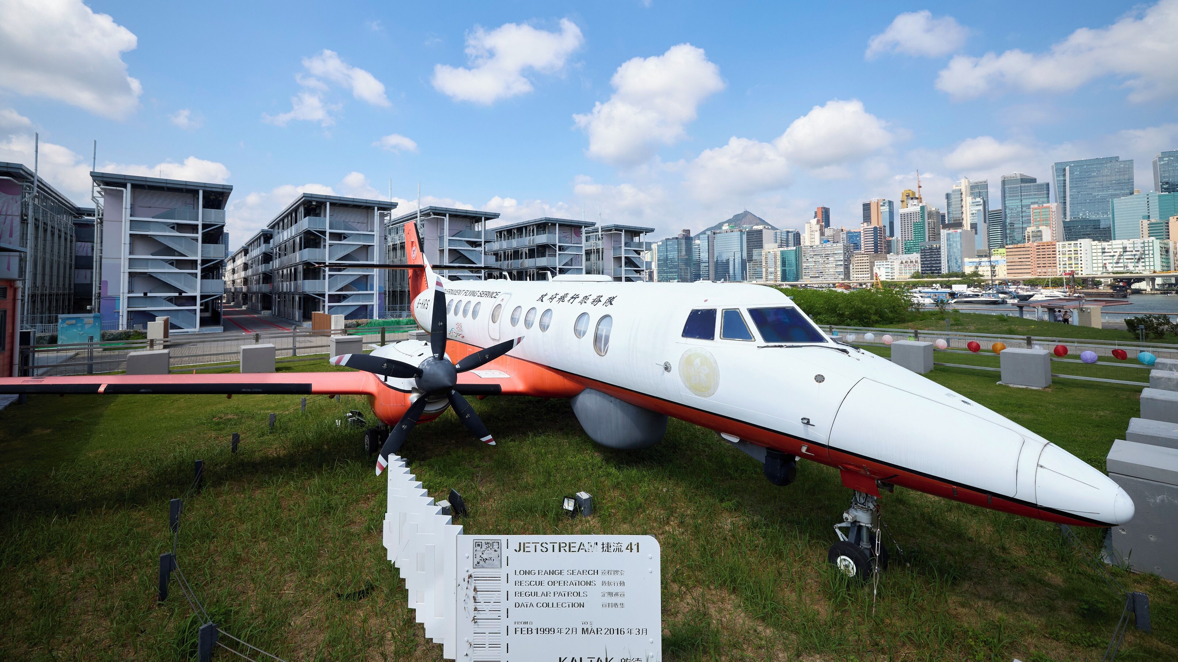 A stationary airplane with a sign detailing its service history, set against a cityscape with modern buildings.