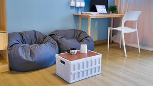 A room with a bean bag, a white crate, a desk with a laptop, a chair, and a window with curtains. - Runway 1331 - Enlight Youth Hostel (Kowloon)
