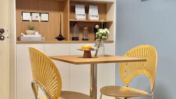 A dining area with a wooden table and two wicker chairs, a cabinet with coffee bags, and a framed abstract painting on the wall.
